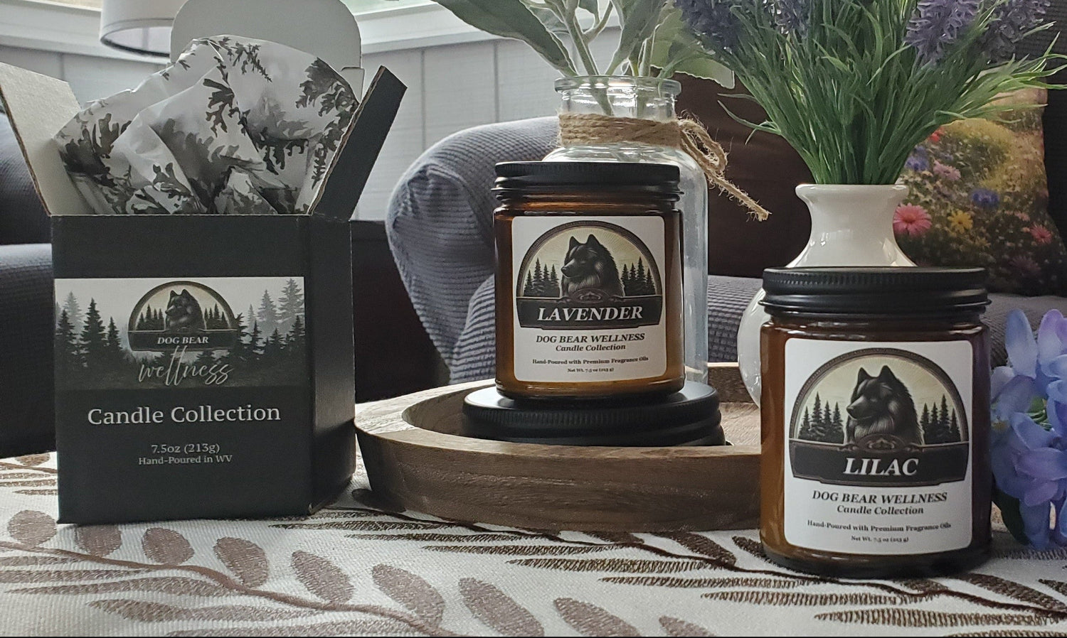 Two scented candles in dark glass jars with black lids are displayed on a table in a cozy living room setting. One jar is labeled 'LAVENDER' and the other 'LILAC', both with matching black gift boxes from the 'Candle Collection'.
