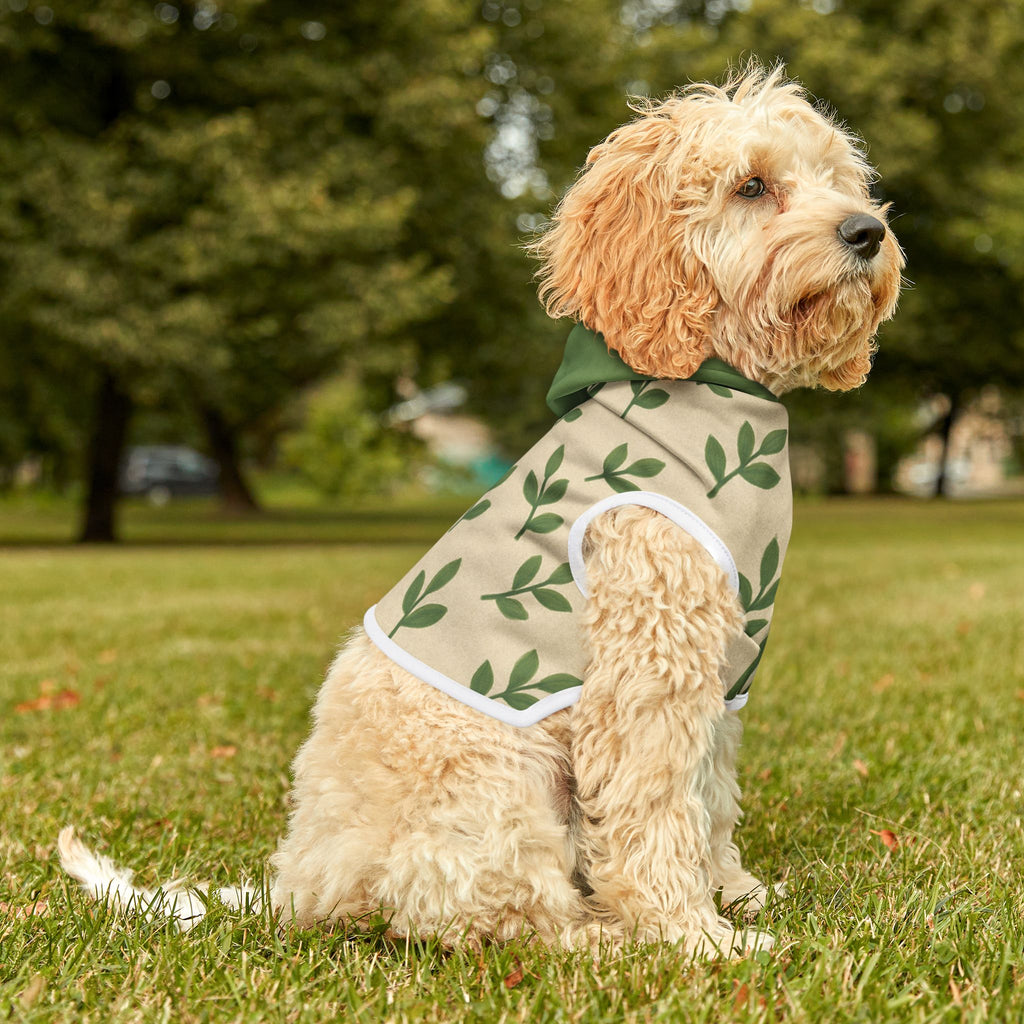 Dog wearing a green and beige patterned coat sitting on grass with trees in the background