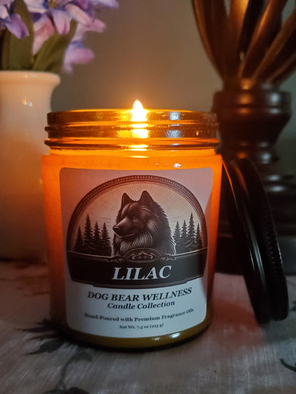 A lit candle in a clear glass jar with a white label reading 'LILAC' and 'DOG HEAD WOLFNESS Candle Collection,' shown on a patterned fabric surface with warm ambient lighting.