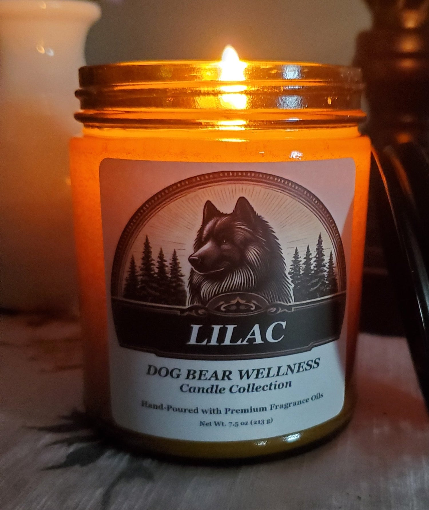 A lit candle in a clear glass jar with a white label reading 'LILAC' and 'DOG BEAR WELLNESS Candle Collection,' shown on a patterned fabric surface with warm ambient lighting.