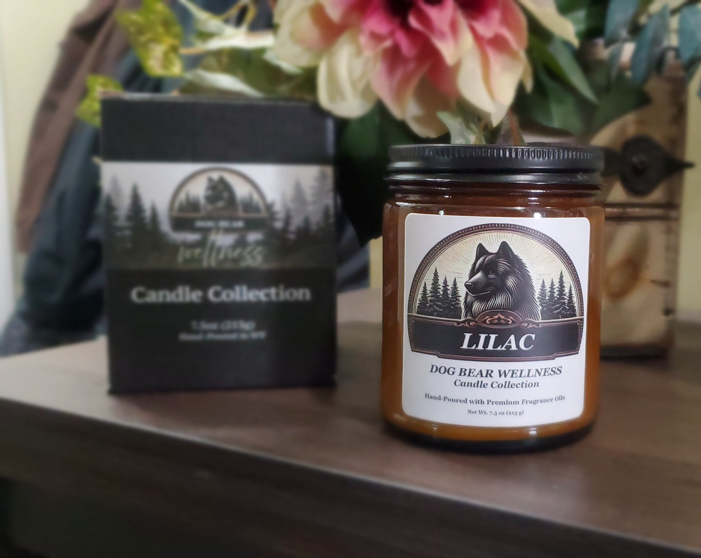 A jar candle labeled 'LILAC Dog Bear Wellness Candle Collection' stands on a wooden surface next to its black retail box, with flowers and a rustic background visible.