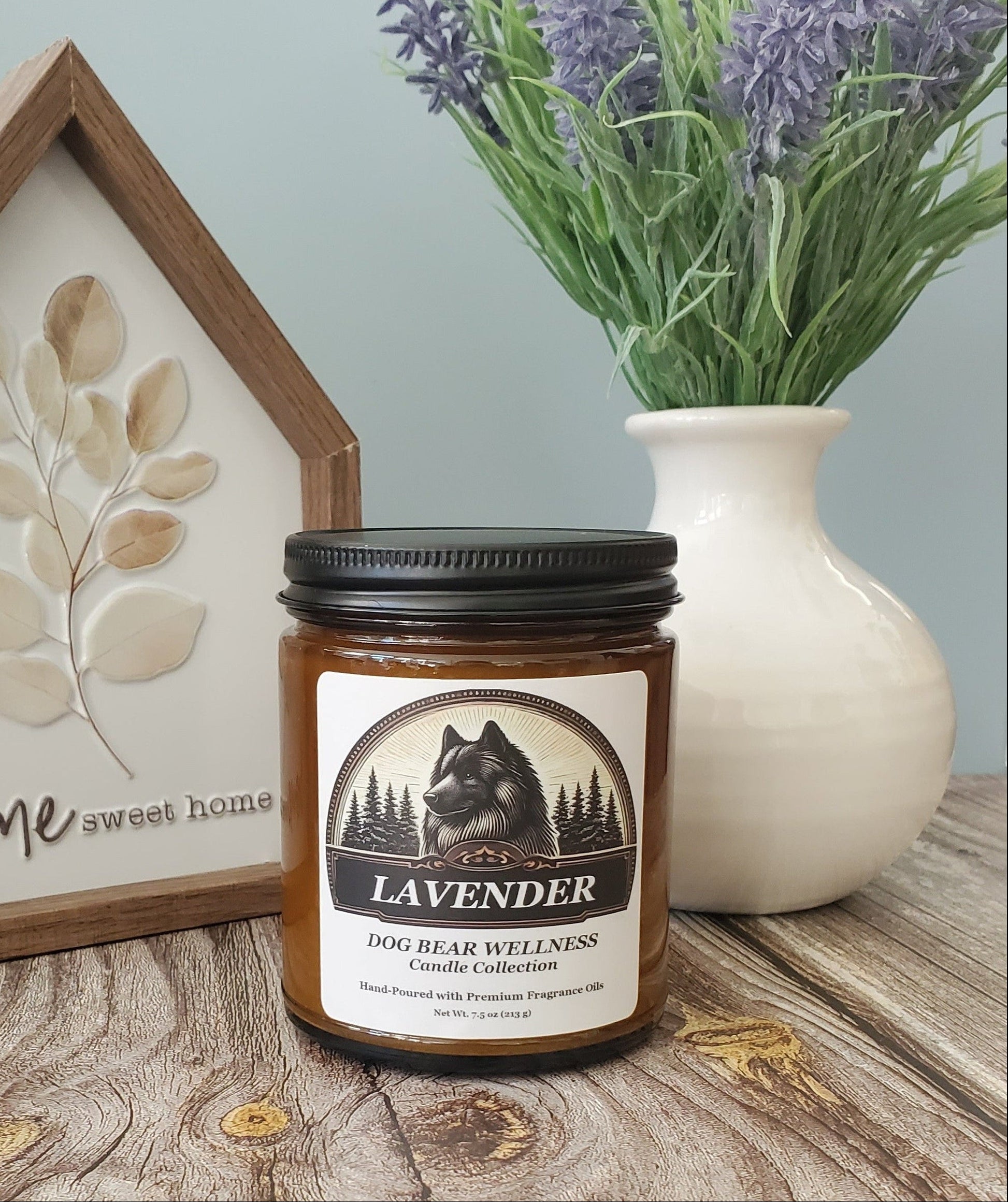 A jar candle labeled 'Dog Bear Wellness Candle Collection Lavender' sits on a rustic wooden surface, styled with a house-shaped decorative sign and a white vase of lavender flowers against a pale blue wall.