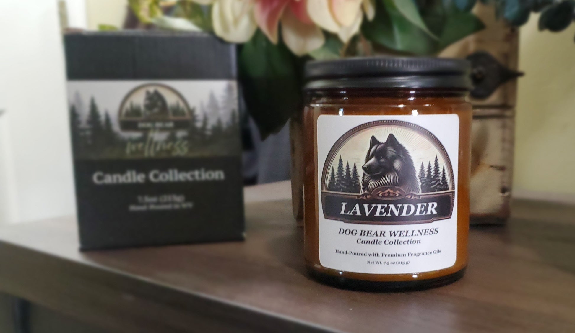 A jar candle labeled 'Lavender Dog Bear Wellness Candle Collection' sits on a wooden surface next to its black product box, with flowers in the background.