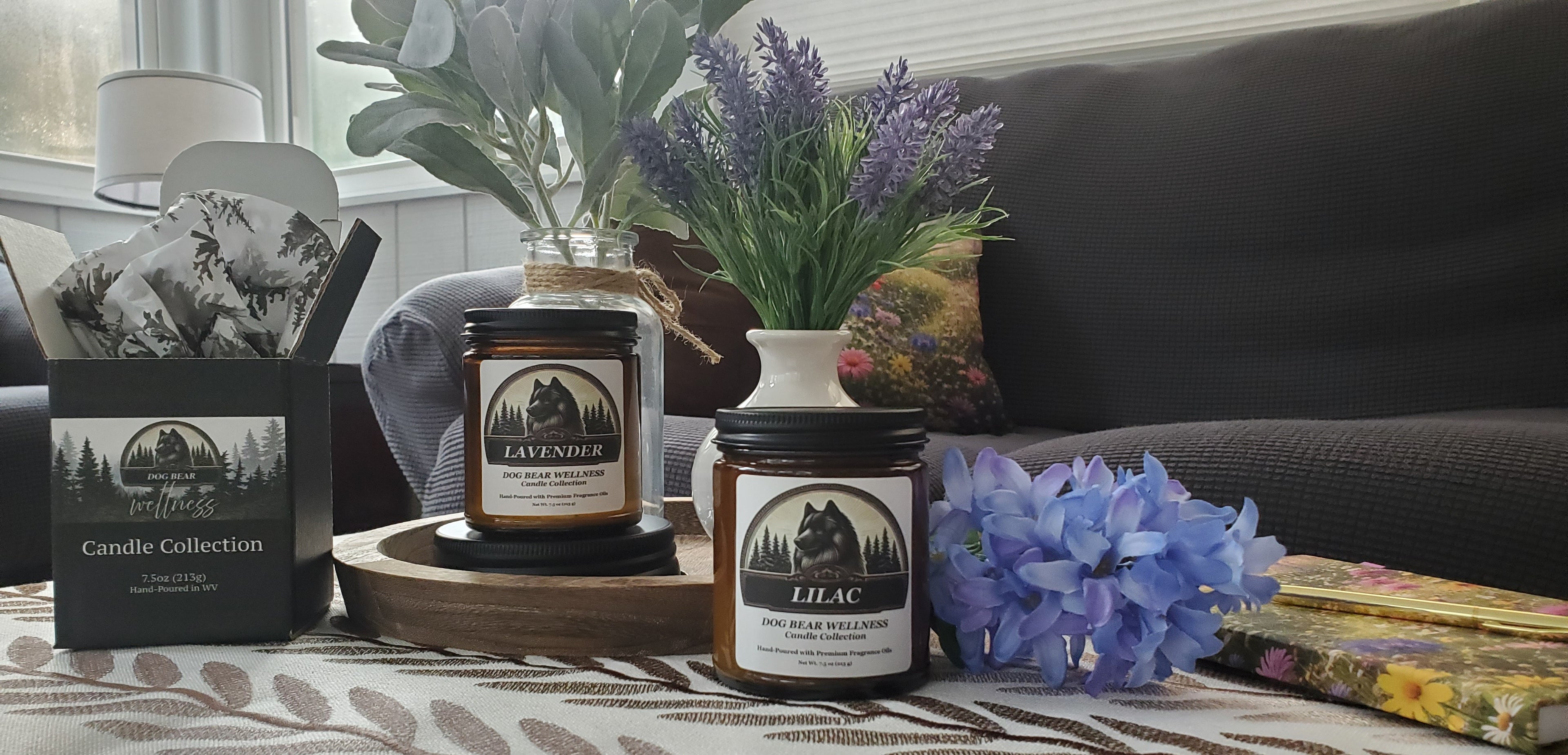 Two scented candles in dark glass jars with black lids are displayed on a table in a cozy living room setting. One jar is labeled 'LAVENDER' and the other 'LILAC', both with matching black gift boxes from the 'Candle Collection'.