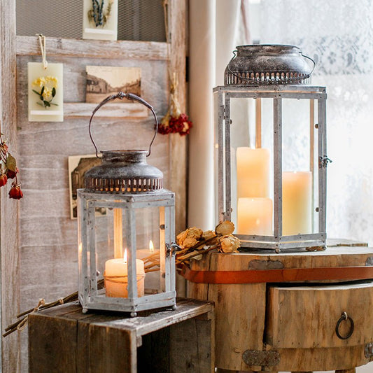Two decorative metal lanterns with glass panels holding lit candles, styled on rustic wooden surfaces in a cozy interior setting.