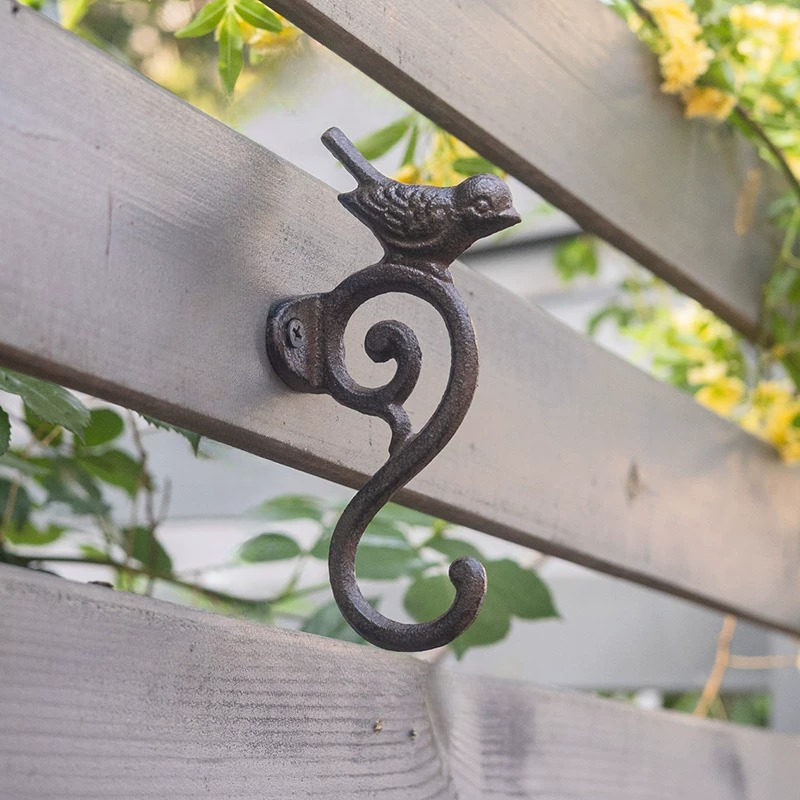 A dark cast iron bird perched on a scroll bracket attached to a wooden fence, with blurred green foliage and yellow flowers in the background.