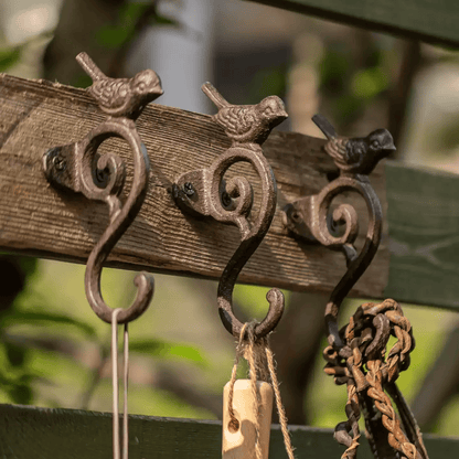 Three rustic cast iron bird-shaped hooks mounted on a weathered wooden fence, with small hanging items including a wooden tag and a braided cord in an outdoor garden setting.
