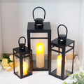 Set of black lanterns with lit candles on a white surface with flowers and greenery.