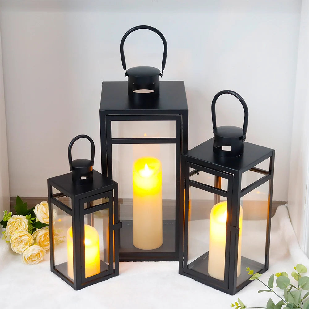 Set of black lanterns with lit candles on a white surface with flowers and greenery.