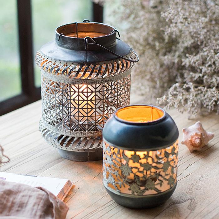 Two decorative metal lantern candles with intricate cutout patterns, lit with warm yellow-orange flames, displayed on a wooden tabletop with a shell and dried flowers in the background.